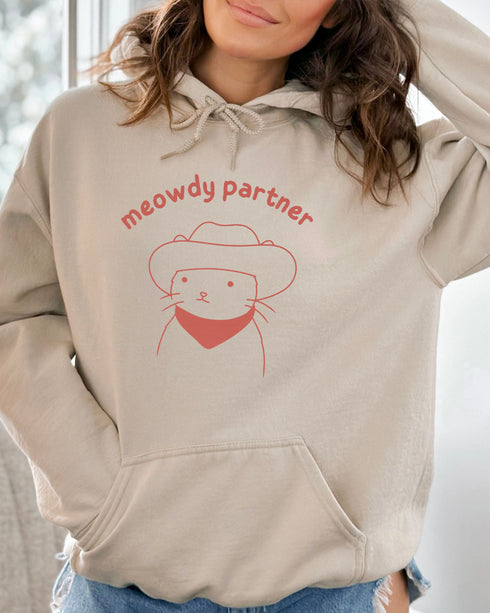Woman wearing a sand beige hoodie featuring a funny retro cowboy cat line-art graphic with the quote 'Meowdy Partner'.